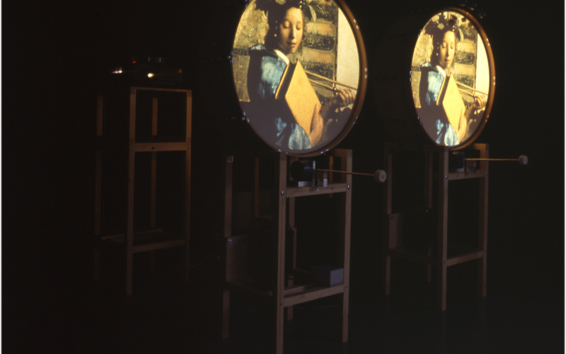 There are three racks to be seen. On two of them round surfaces are fixed. On each of them the same image is projected: a young woman with a large book in her hand.