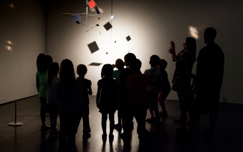 Kids are standing in the dark museum, gazing at an mobile, that is illuminated