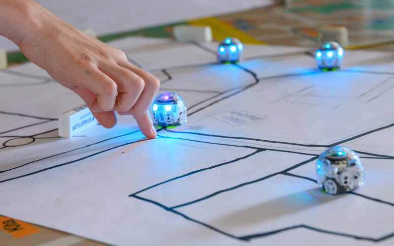 On the photo: Small, blue illuminated robots drive along black lines on a sheet of paper.