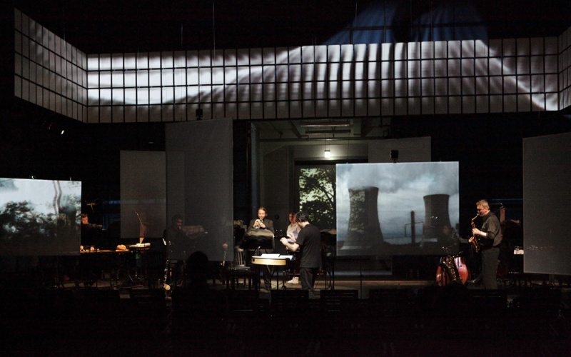 A music ensemble on a dusky stage. In the background a picture of a nuclear power station