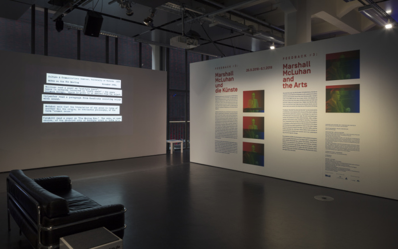 In the foreground on the left side is a black sofa. At the back left there is a partition wall with text projection. In the back on the right there is a wall printed with text and pictures.