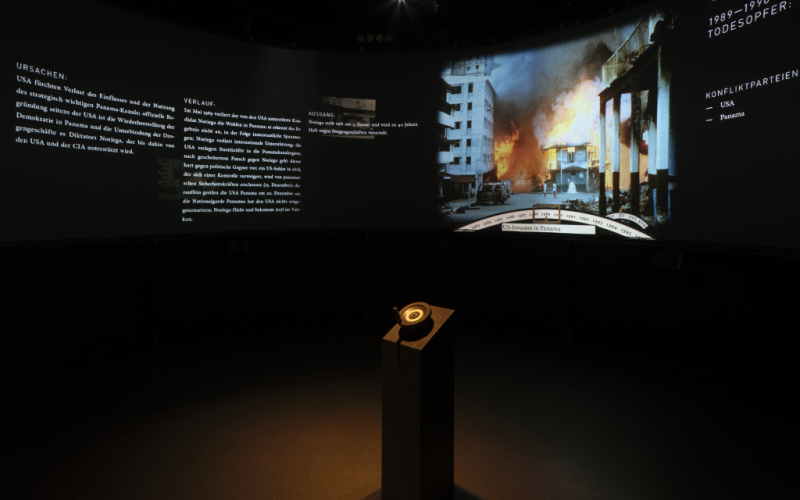 Das Foto zeigt einen verdunkelten Raum, der auf einer großen Panoramaleinwand Kriegsszenarien präsentiert und mit Text ihre Ursachen aufzeigt.