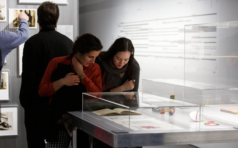 Zu sehen sind zwei Frauen, die sich über eine Vitrine in der Ausstellng beugen und das ausgestellte Buch genauer betrachten.