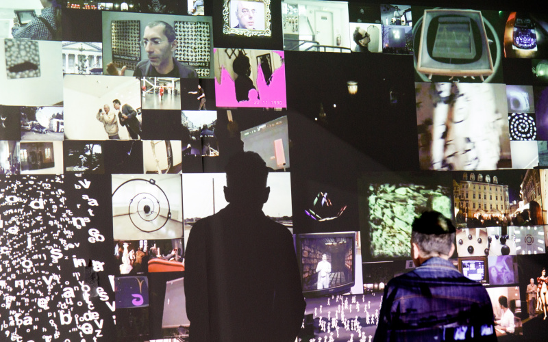 You can see a video installation. Many smaller images are projected onto a wall, creating a mosaic-like image. In front of it stands a man. He casts a conspicuous shadow on the wall.