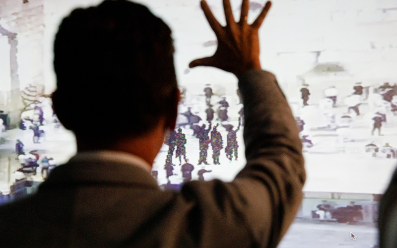 A picture can be seen on a wall on which shadowy figures raise their hands to salute. In front of it stands a man, his back to the observer and also raising his hand.