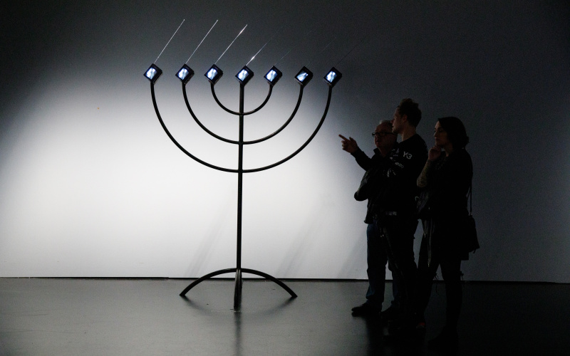 You can see a large, black Menorah with seven small screens with right-facing antennas instead of candles. On the right there are three people looking at it. One of them points to the installation.