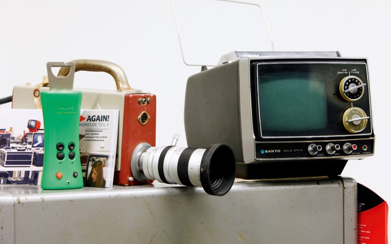 Ein sehr alter, kleiner TV-Monitor, eine alte Videokamera und eine Fernbedienung mit integriertem Flaschenöffner 