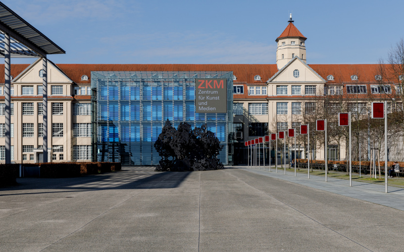 The ZKM from the outside. You can see frontally the cube with the total glazing and the sound pavilion in front of it, the long industrial building of the ZKM as well as the square of the human rights with the several electronic flags standing in a row