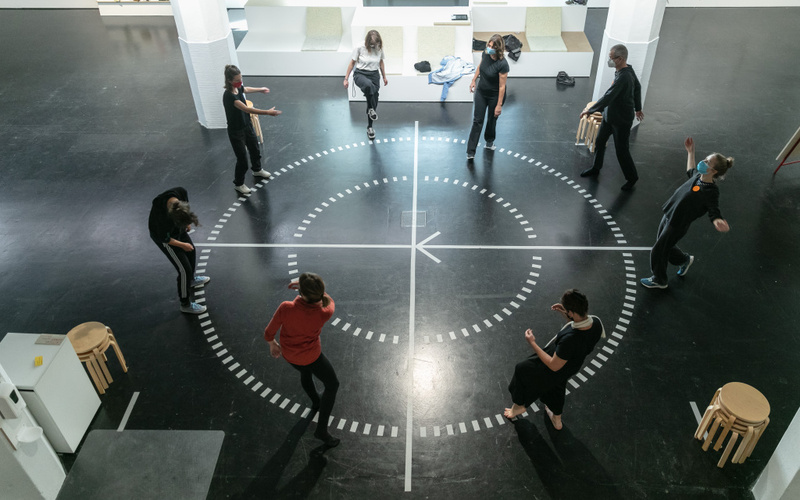 A group of people stands in a circle on a round diagram on a hall floor. The people seem to be in motion.
