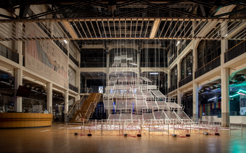 Installation view of Chiharu Shiota`s work "Connected to Life". You can see bedsteads lined up and connected to each other, crisscrossed by red tubes that rise further and further from the floor and together form a kind of pathway upwards. 