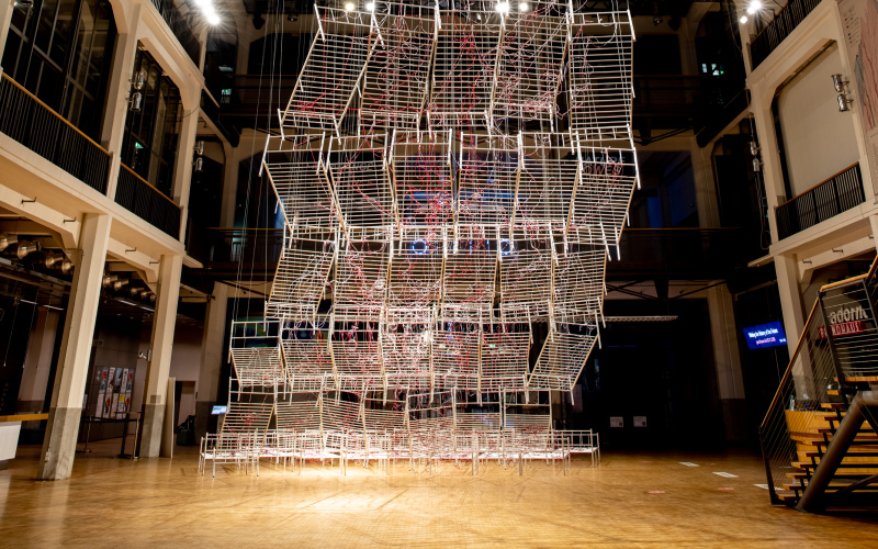 Installationsansicht von Chiharu Shiota`s Werk »Connected to Life«. Zu sehen sind aneinandergereihte, miteinander verbundene Bettgestelle, durchzogen von roten Schläuchen, die sich immer weiter vom Boden abheben und gemeinsam eine Art Weg aufwärts bilden.
