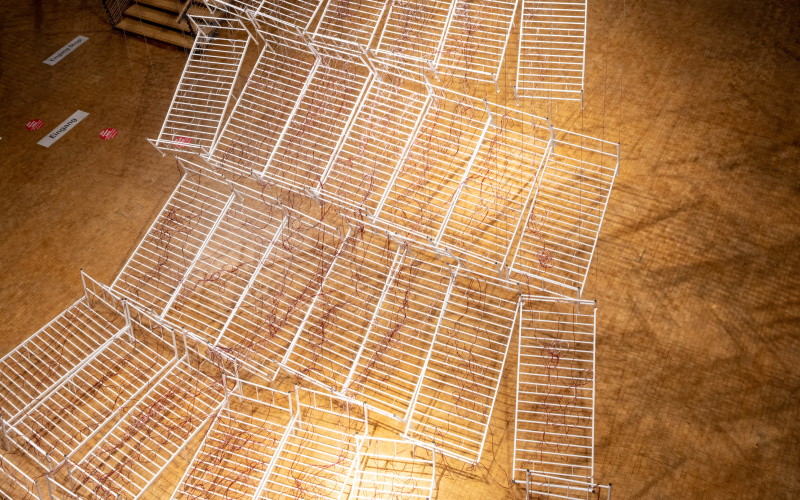Installationsansicht von Chiharu Shiota`s Werk »Connected to Life«. Zu sehen sind aneinandergereihte, miteinander verbundene Bettgestelle, durchzogen von roten Schläuchen, die sich immer weiter vom Boden abheben und gemeinsam eine Art Weg aufwärts bilden.