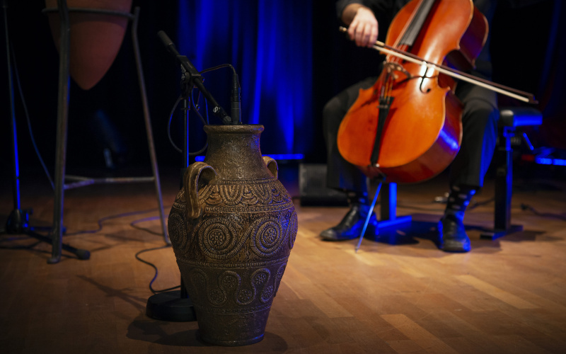 Zu sehen ist eine braune, verzierte Vase im Zentrum und rechts dahinter ein Person am Cello