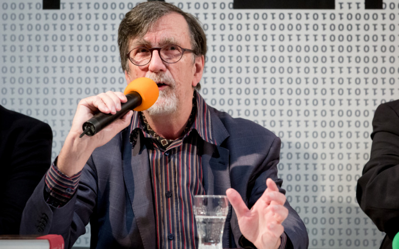Bruno Latour during the press conference for the exhibition "Reset Modernity" 2016. He holds a microphone in his right hand and makes an open, explanatory gesture with his left hand. A glass of water is in front of him.