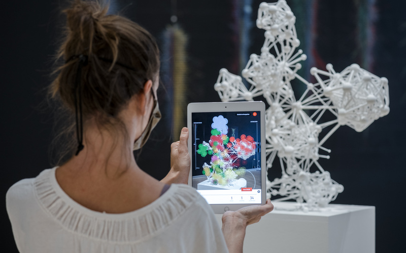 Eine Person steht vor einer weißen 3D-Netzwerk-Skulptur und hält ein Tablet hoch, auf dem mittels Augmented Reality die Skulptur näher untersucht werden kann.