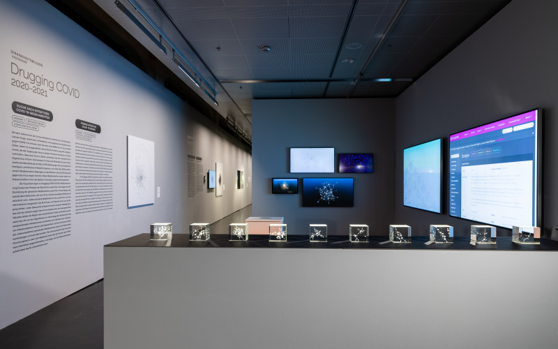 An exhibition space. In the foreground are 10 small glass boxes showing fine networks. In the background hang 4 screens that also show networks. On the right hang two large screens, on the left is an explanatory text on the wall.