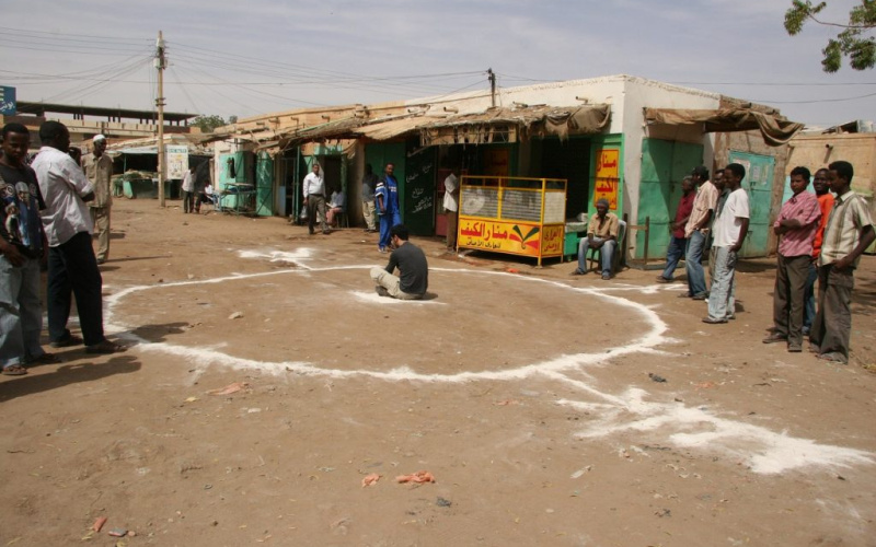 Künstler Barış Seyitvan sitzt innerhalb eines auf dem Boden aufgemalten weißen Kreises in einem sudanesischen Dorf.