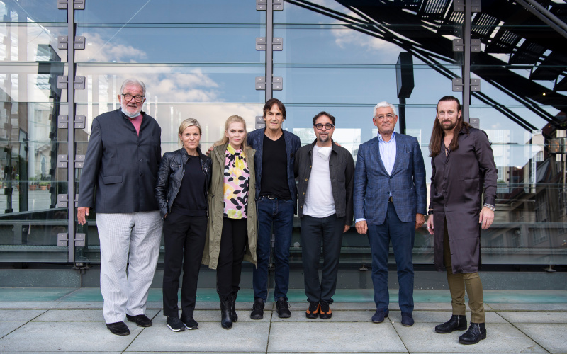 Seven people stand in front of the glass wall of the ZKM cube and look into the camera.