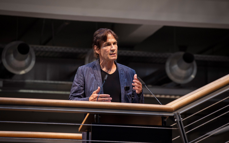 The artist Jean-Remy stands on the ZKM foyer stairs at the railing behind a lectern and speaks. His gestures accompany his speaking.