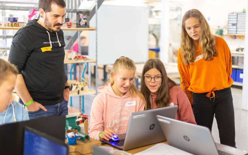 Ein Foto der Veranstaltung Hack to the Future, zwei junge Teilnehmerinnen schauen auf ihre Laptops, daneben stehen eine Mentorin und ein Mentor.