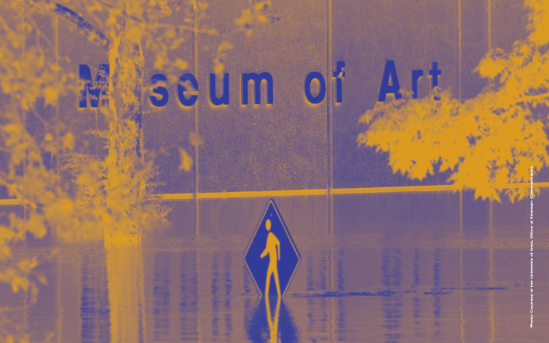 Colour-distorted photo of a flooded street. A street sign with a running person on it rises out of the water. The building in the background reads "Museum of Art".