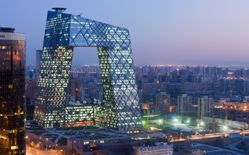 The picture shows a cityscape, at dusk. The center is a large illuminated building in the form of a gate or arch. 