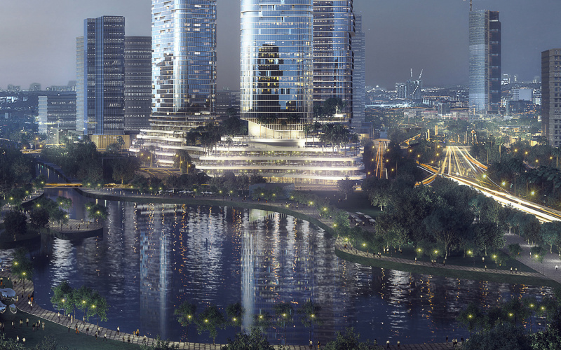 The photo shows a large modern skyscraper in the center, in front of other skyscrapers. In front of it is water, in which all the lights and buildings are reflected. The photo was taken in the dark and the whole city is lit.