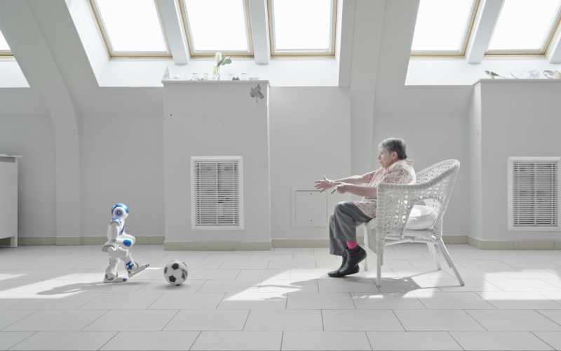 A small humanoid robot plays soccer with a senior citizen in a nursing home.
