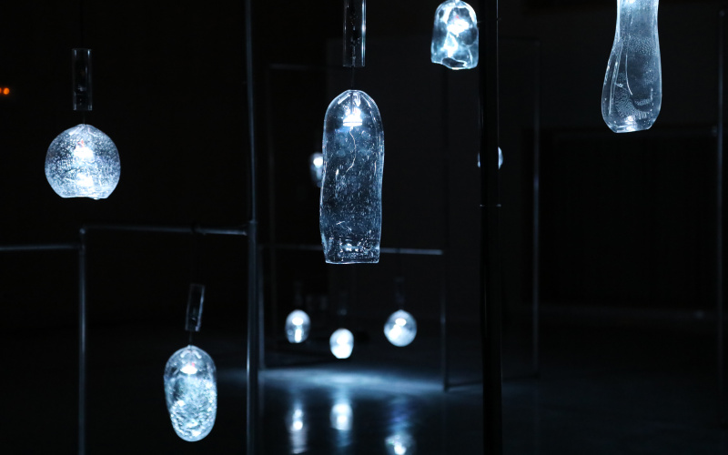 Glass vessels of various shapes hang in a dark room and glow dimly.