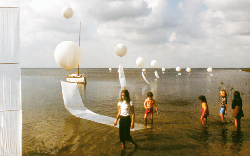 Zu sehen sind fünf Kinder, die durch Wasser laufen. Auf der linken Seite sind weiße Luftballons zu sehen.