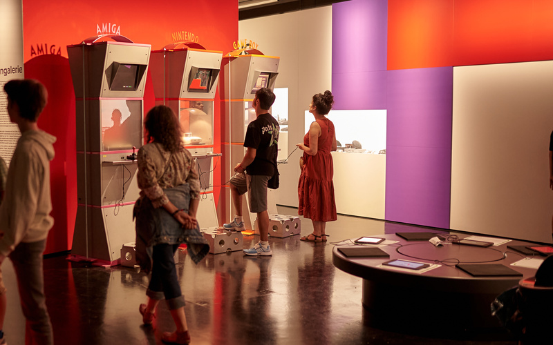 Ausstellungsansicht zkm_gameplay. the next level. Mehrere Personen stehen vor verschiedenen Spielautomaten, die vor einer roten Wand stehen.