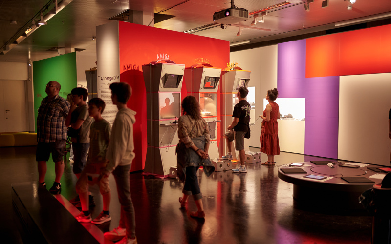 Ausstellungsansicht zkm_gameplay. the next level. Mehrere Personen stehen vor verschiedenen Spielautomaten, die vor einer roten Wand stehen.