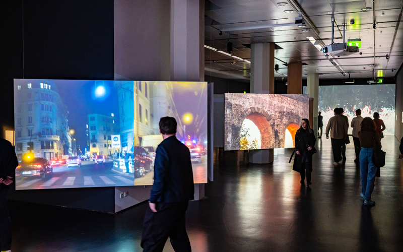 Videos showing views of Rome are projected onto three screens distributed around the room.