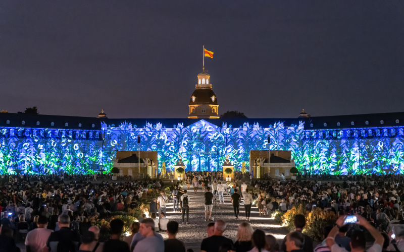 Karlsruhe Castle illuminates in a blue-green plant pattern. You can also see the many people sitting in the audience.