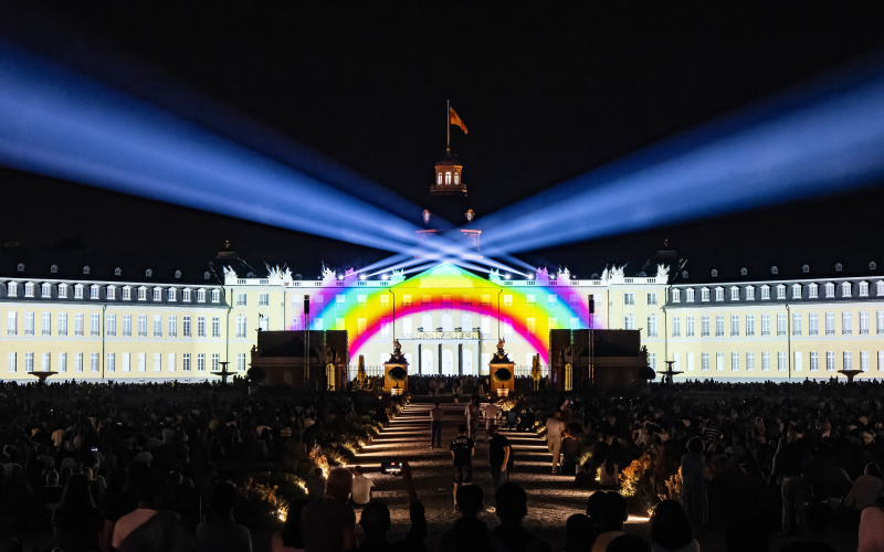 You can see the facade of the Karlsruhe Castle. Projected on it is a rainbow with spotlights shining in the black sky.