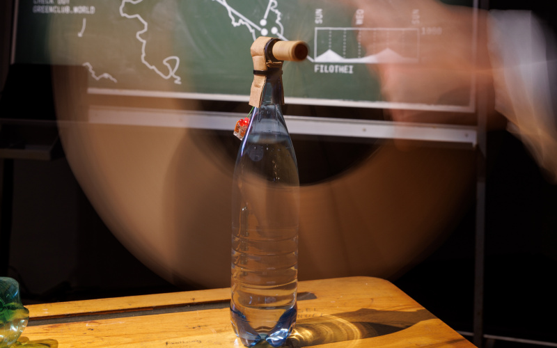 In the foreground there is a glass bottle on which a kind of tube was fixed. In the background there is a board with statistics.