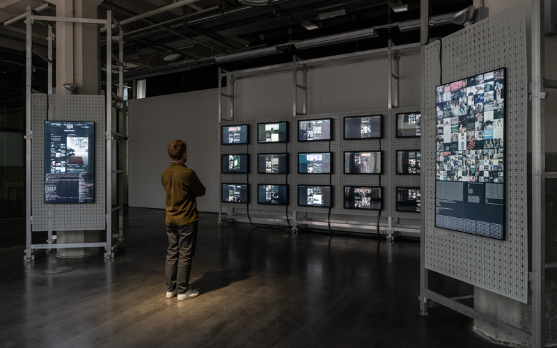 You can see a large open space with a man standing in the middle looking at many screens. The screens hang on the wall and display many different codes and shapes. 
