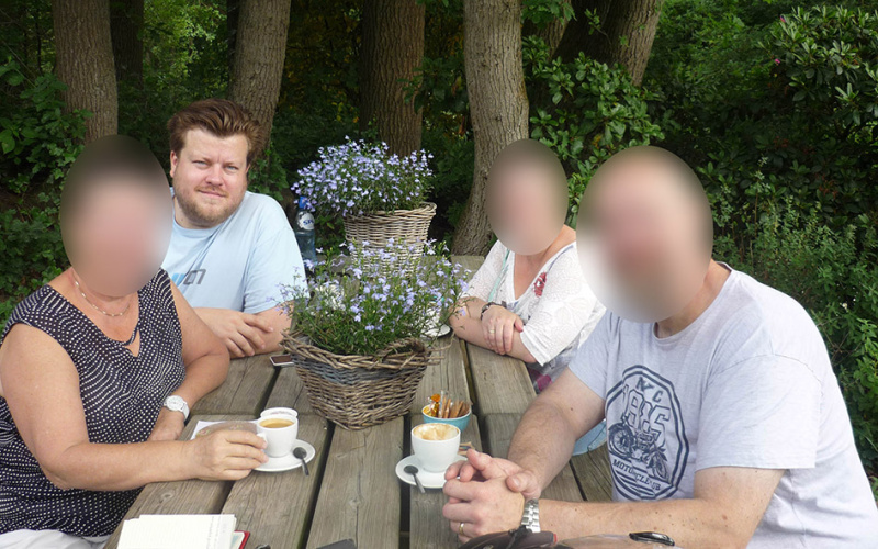 Auf dem Foto sieht man den Künstler Constant Dullaart, der mit drei zensierten Personen an einem Holztisch im Freien sitzt.