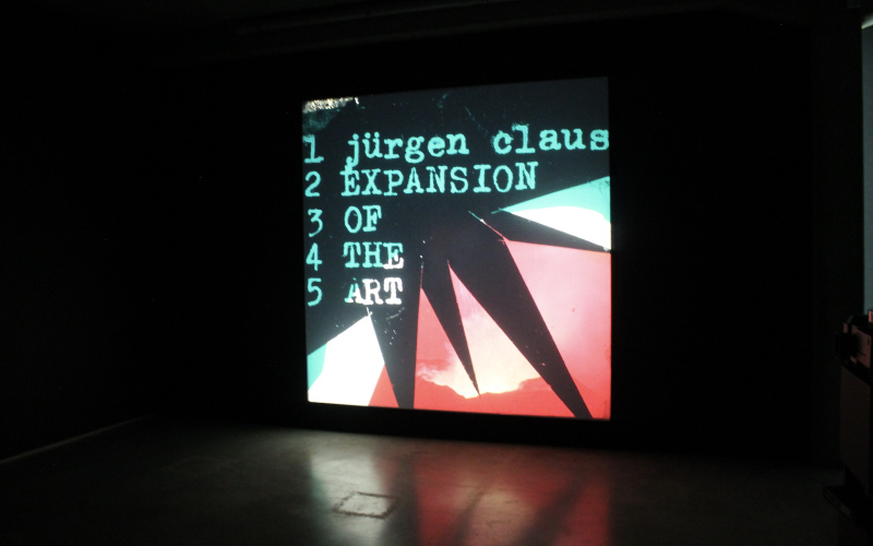Foto einer Multimediaprojektion, ein Bild in Rosa, Schwarz und Türkis mit geometrischen Formen. Darauf der Text: »Jürgen Claus, Expansion of the Art«
