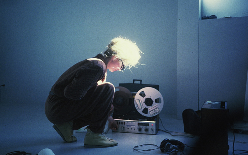 In the picture, a woman is sitting in a crouching position and adjusting something on a receiver that is standing diagonally next to her. Around her are some technical objects on the floor and she herself has sunglasses on and is illuminated through a win