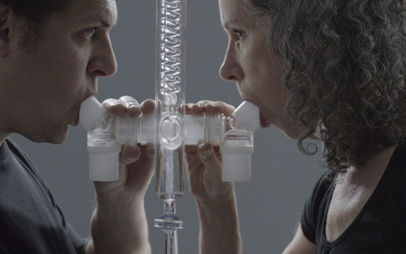 In the picture, there are two people blowing into a tube construction, one on the left and one on the right.