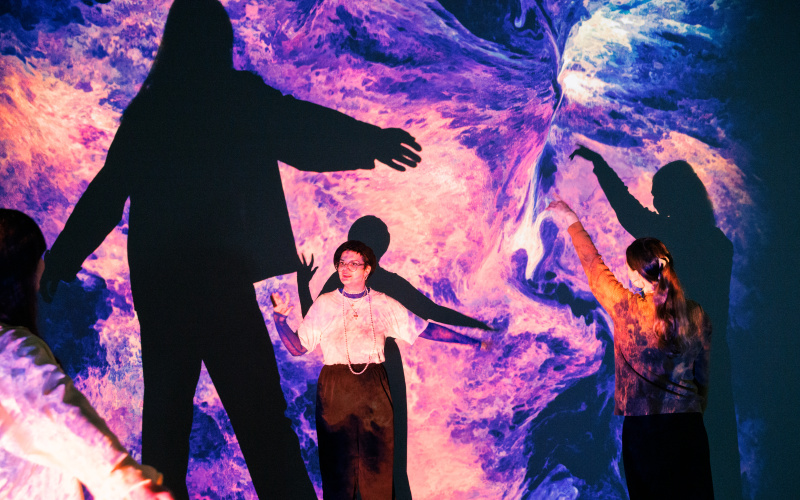 On display is a large canvas, which is covered with an organic, blue and pink pattern. In front of it are three people stretching their arms upwards.