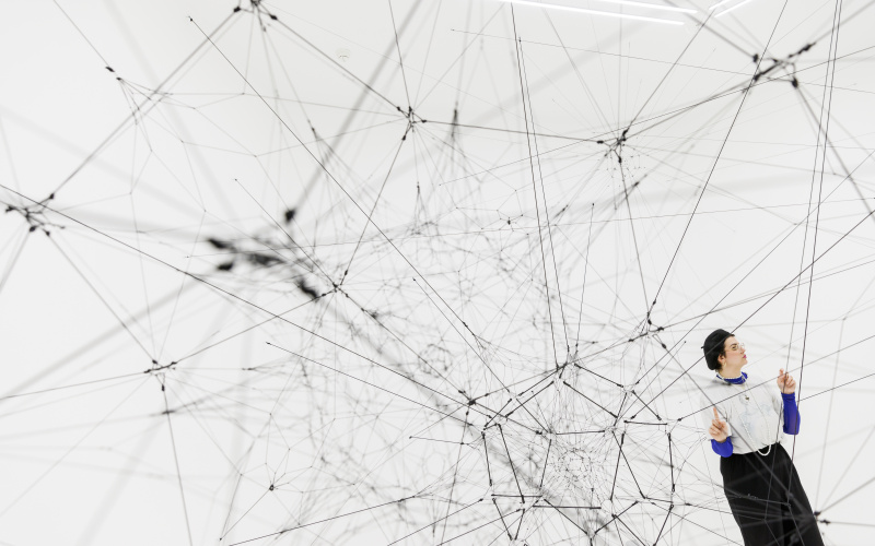 You can see a large net of threads in a white room. On the right side there is a person touching one of the threads.