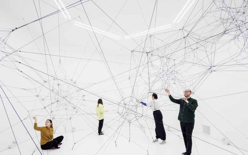 You can see a large net of threads in a white room. Several people are standing in this space and touching these threads.