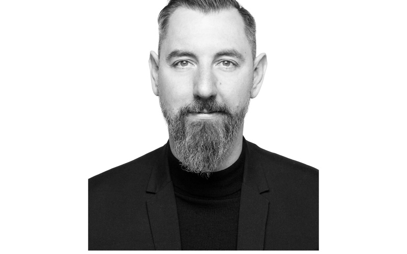 Zu sehen ist ein Porträt von Stephan Schwingeler. es ist ein schwarz weiß Foto und zeigt ihn ab der brust aufwärts. Er trägt einen Bart und ein schwarzes T-shirt und schaut frontal in die Kamera.