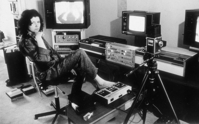 Das Foto ist schwarz-weiß und zeigt Ulrike Rosenbach, die Medienkünstlerin, in ihrem Studio in den 70er Jahren. Ulrike Rosenbach sitzt auf einem Drehstuhl und ist umgeben von Kameraequipment und Röhrenbildschirmen. 