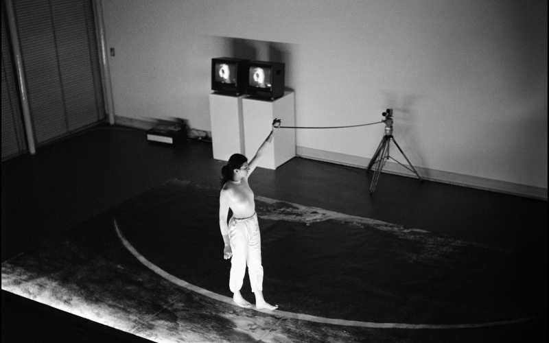 Ulrike Rosenbach, »Die einsame Spaziergängerin«, 1979. Zu sehen ist eine Person auf einer Bühne, die barfüßig auf einer gebogenen Linie läuft. Dabei hält sie ein Seil in der Hand, das an einem Stativ befestigt ist. Das Bild ist schwarz-weiß. 