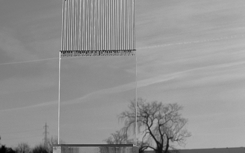 In the middle of a landscape stands a large glass sculpture. The lower part of the sculpture is made of transparent glass and the upper part is made of glass, which looks like a harp and has no clear edge upwards. The image is black and white.
