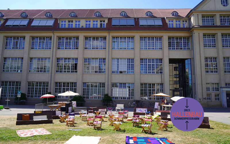 A meadow with various seating options can be seen. In the background is the Hallenbau.