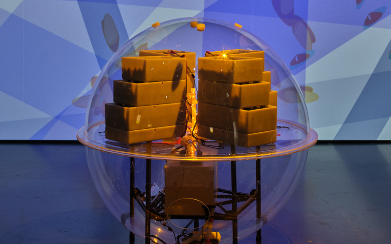 »Active Living Infrastructure: Controlled Environment (ALICE) 2.0« von Julie Freeman. In einer durchsichtigen Kugel befinden sich rechteckige Barren, in deren Mitte befindet sich ein Licht. Die Kugel steht vor einem großen Bildschirm.